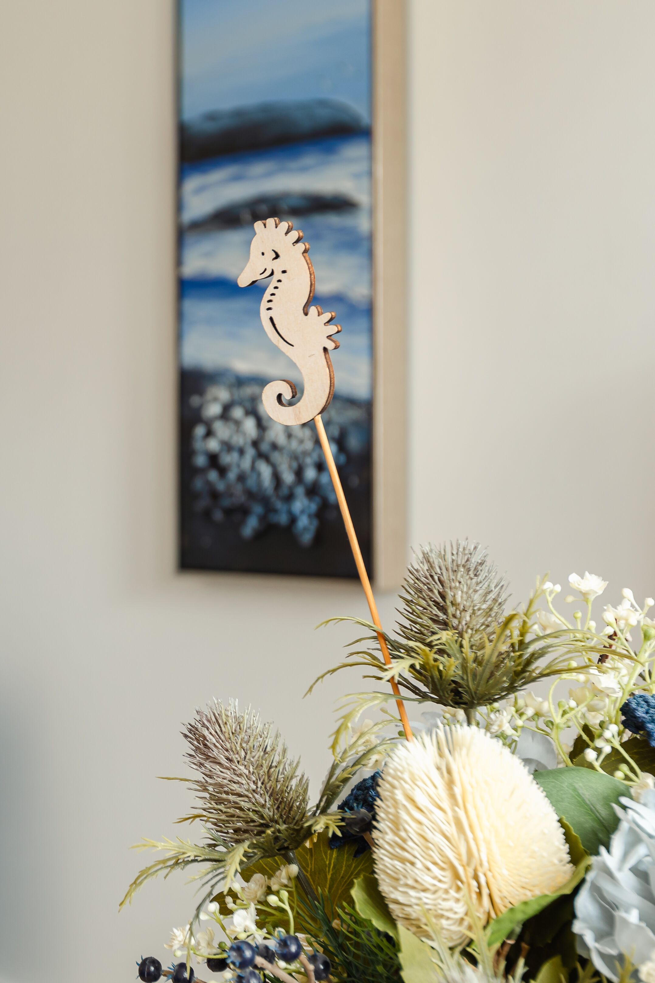 A wooden seahorse decoration on a stick, a framed painting of a seascape, and a floral arrangement with various plants and flowers.