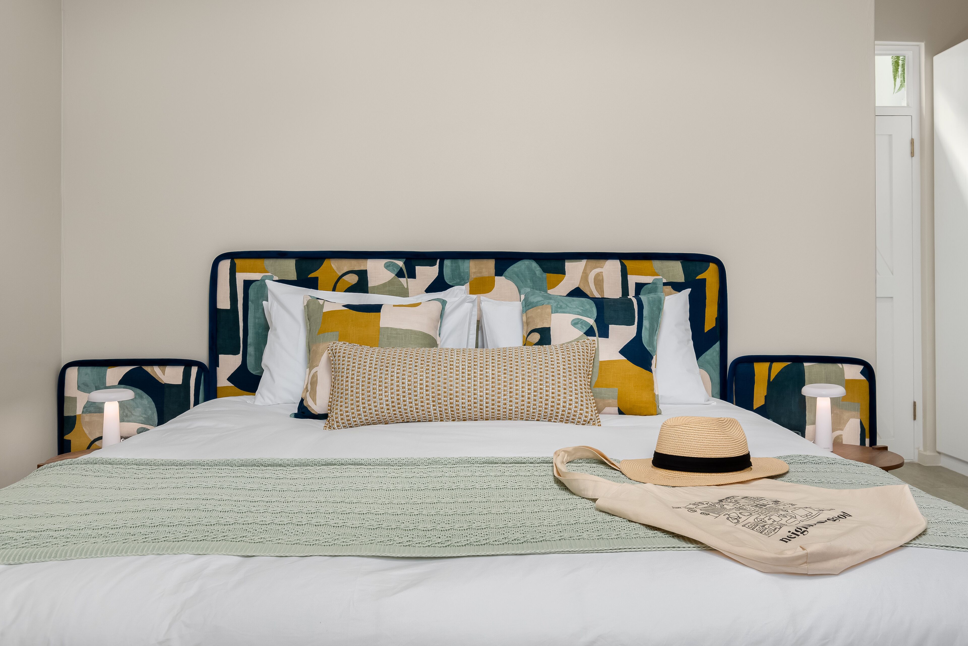 A neatly made bed with a patterned headboard, a green knitted throw, and a beige hat on the pillows.