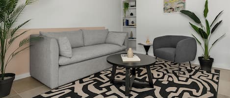 A modern living room with a grey sofa, a round black table, and a grey armchair. There are two potted plants, one large and one small, and a wall-mounted shelf with books and decorative items.