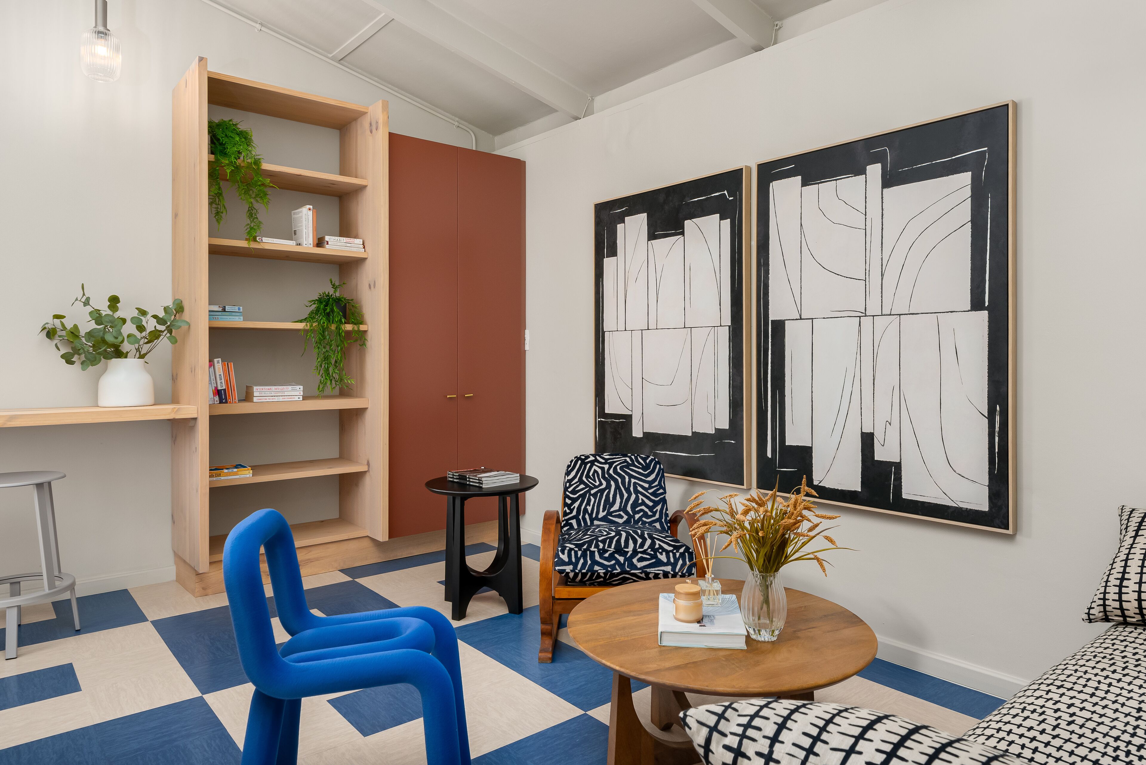 A modern living room with a blue chair, a round wooden table, a bookshelf, and two abstract paintings on the wall.