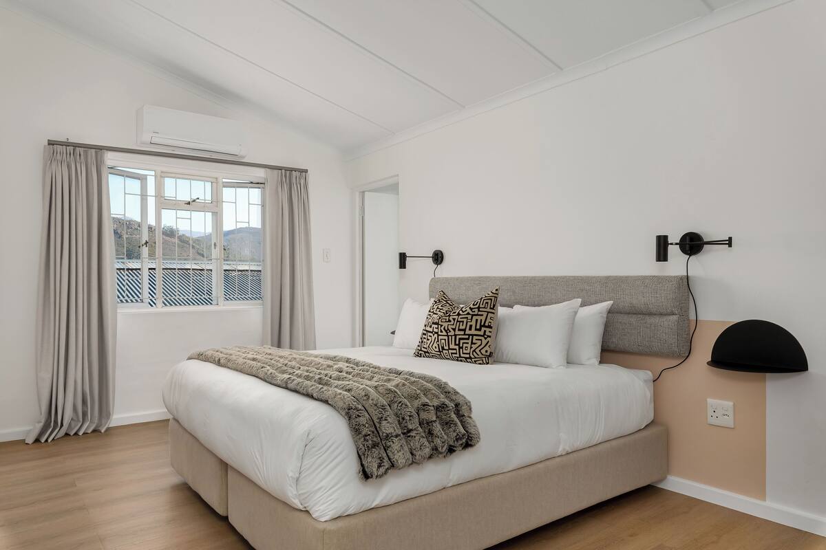 A bedroom with a bed, a window with curtains, an air conditioning unit, and a wall-mounted lamp.
