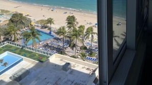 A view from a balcony overlooking a beach with palm trees, a pool, and lounge chairs.