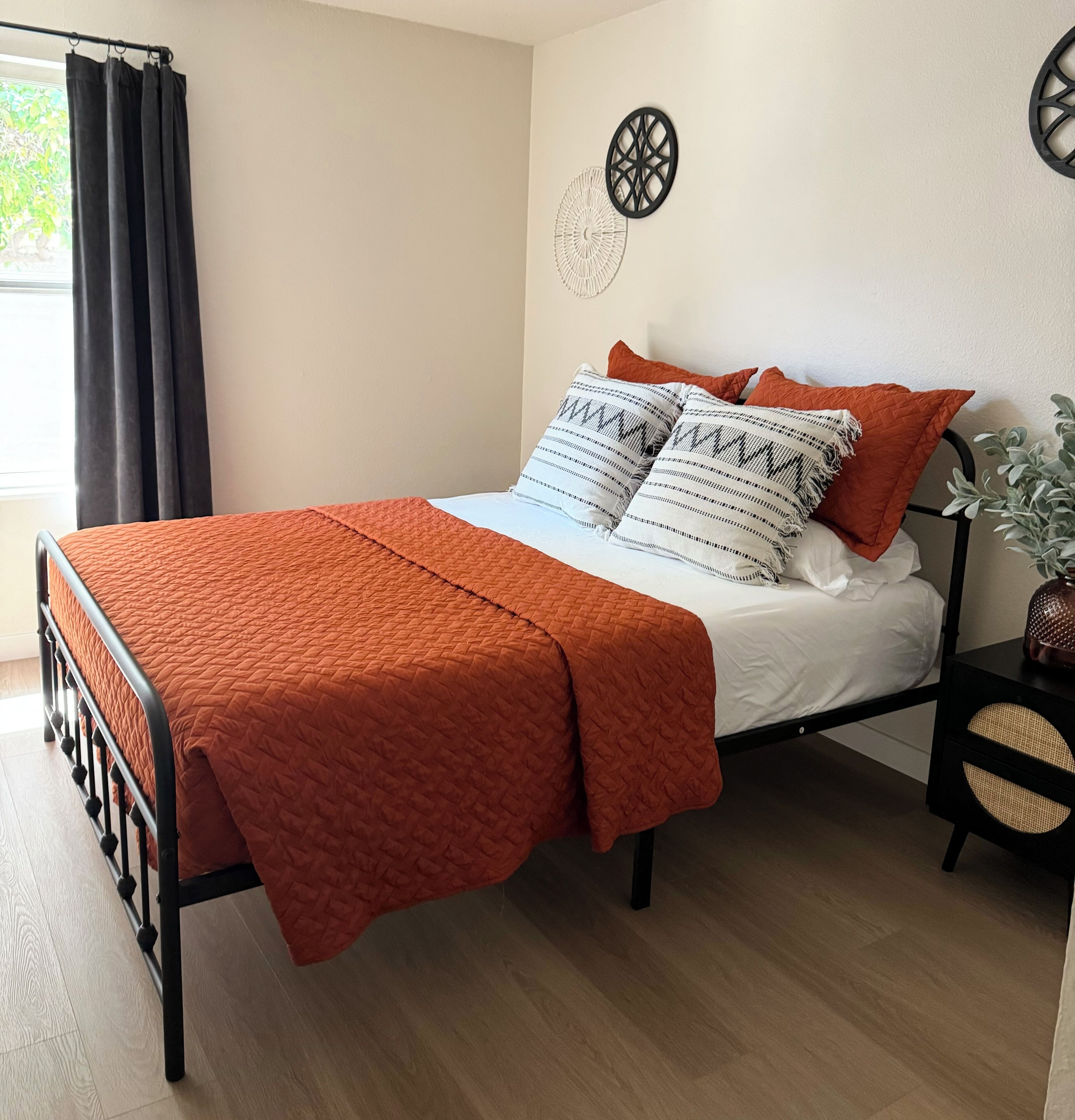 Una cama con colcha naranja, ropa de cama blanca y almohadas estampadas. Tiene un cabecero de metal negro, una mesita de noche con una cesta tejida y una decoración en la pared.