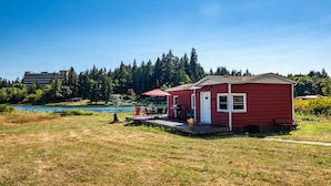A red cabin with a deck, a grill, and a table with chairs, surrounded by a lake and forest. - Peaceful Beachfront Cottage - Large Deck & Fire Pit - Gorgeous Views & Wildlife (Bremerton)
