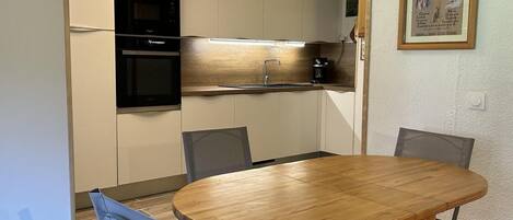 A compact kitchen with a round wooden table and four chairs, a black oven, and a stainless steel sink.