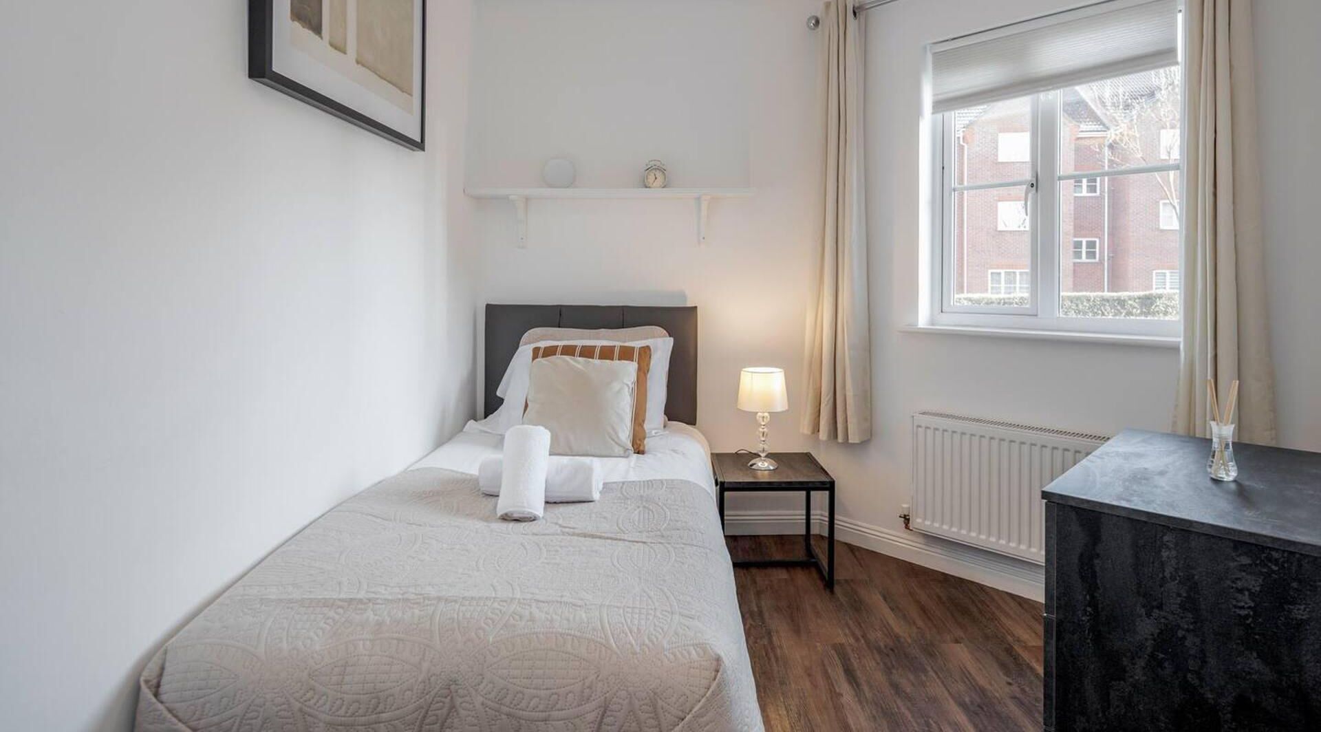 Un dormitorio ordenado con cama, mesita de noche, lámpara y vistas al exterior a través de una ventana.