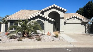 A single-story house with a tiled roof, a garage, and a landscaped front yard with plants. - Lake House Pool & Palms (Goodyear)