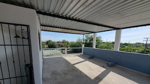 Varanda coberta com cobertura de metal, guarda-corpo de vidro e vista para vegetação e edifícios ao fundo.