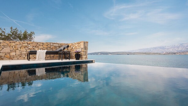 Pared de piedra con una silla de lounge y una mesa junto a una piscina azul cristalina que refleja el cielo.