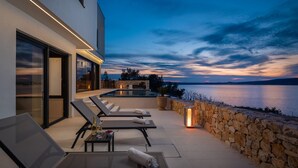 Un patio moderno al aire libre con sillones de descanso, una mesa con bebidas y un muro de piedra con vista a un cuerpo de agua al anochecer.