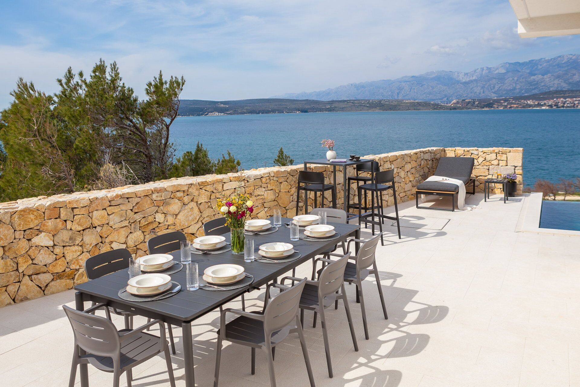 An outdoor dining area with a stone wall, a table set for four, and a view of the sea and mountains.