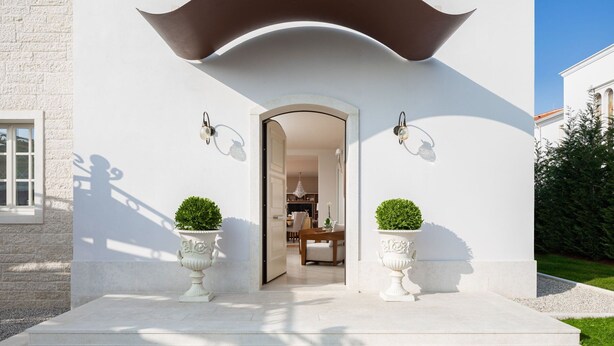 Une maison moderne avec une façade blanche, un toit incurvé et deux plantes en pot placées sur les marches de l’entrée.