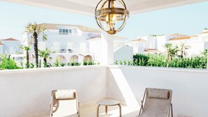 Un balcon avec deux chaises longues et une table, donnant sur un bâtiment blanc orné d’arcades et bordé de palmiers.