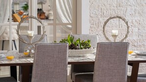 Une table à manger avec quatre chaises grises à revêtement rembourré, deux chandeliers avec des bougies blanches, un bol décoratif contenant des plantes vertes et un verre de jus d’orange posé sur la table.
