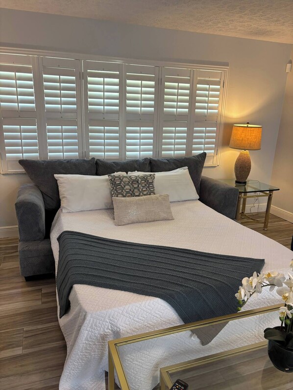 A bedroom with a large bed, a grey sofa, a lamp, and a mirror. - Heated Pool Oasis - Sleeps 12 (Port St. Lucie)