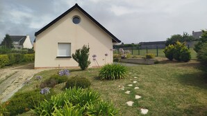 A small, single-story house with a beige exterior, a small window with a shutter, and a circular window above it. There is a garden with various plants and flowers in front of the house. - Maison Plein Pied a Loué a Camaret sur mer (Camaret-sur-Mer)