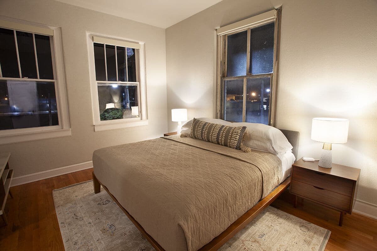 A bedroom with a bed, nightstands, and two windows.