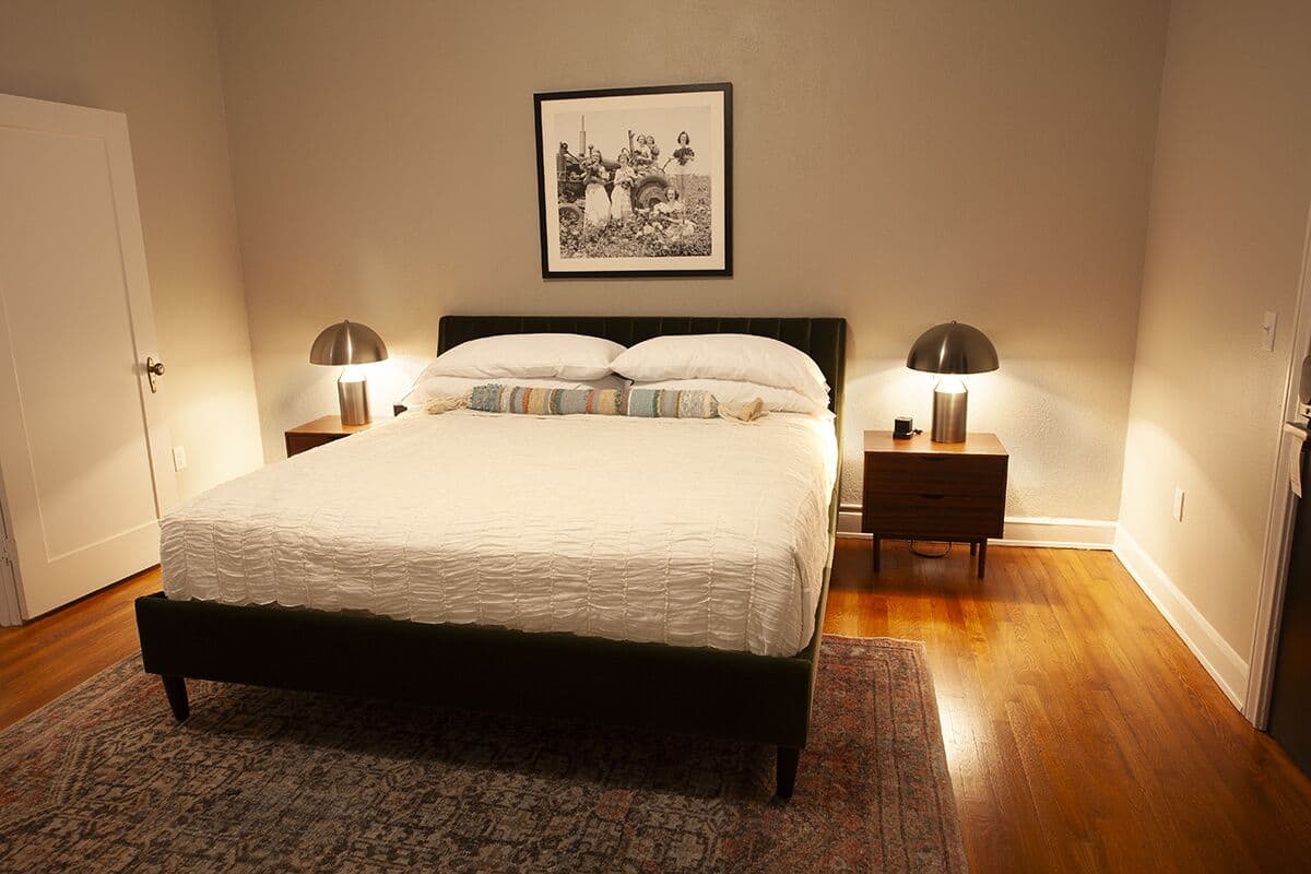 A neatly made bed with a white duvet, two bedside lamps, a framed picture on the wall, and a wooden floor.