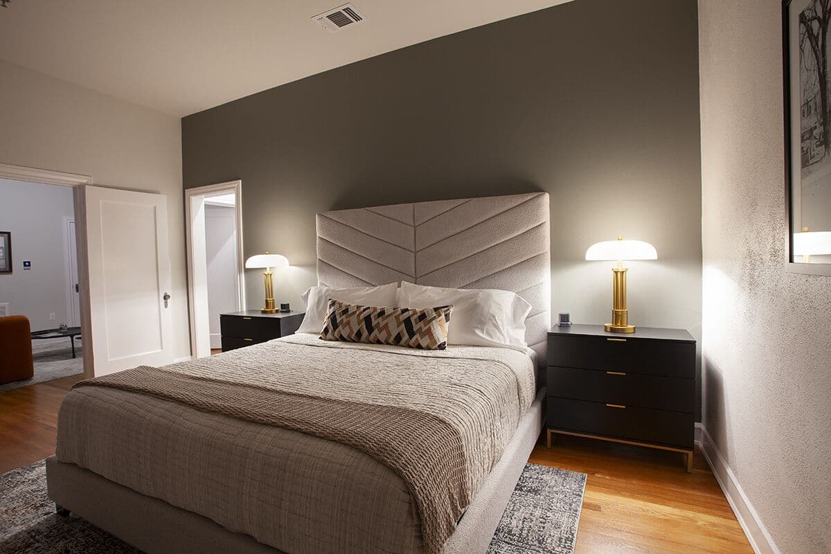 A bedroom with a large bed, two bedside tables with lamps, a dark dresser, and a door leading to another room.