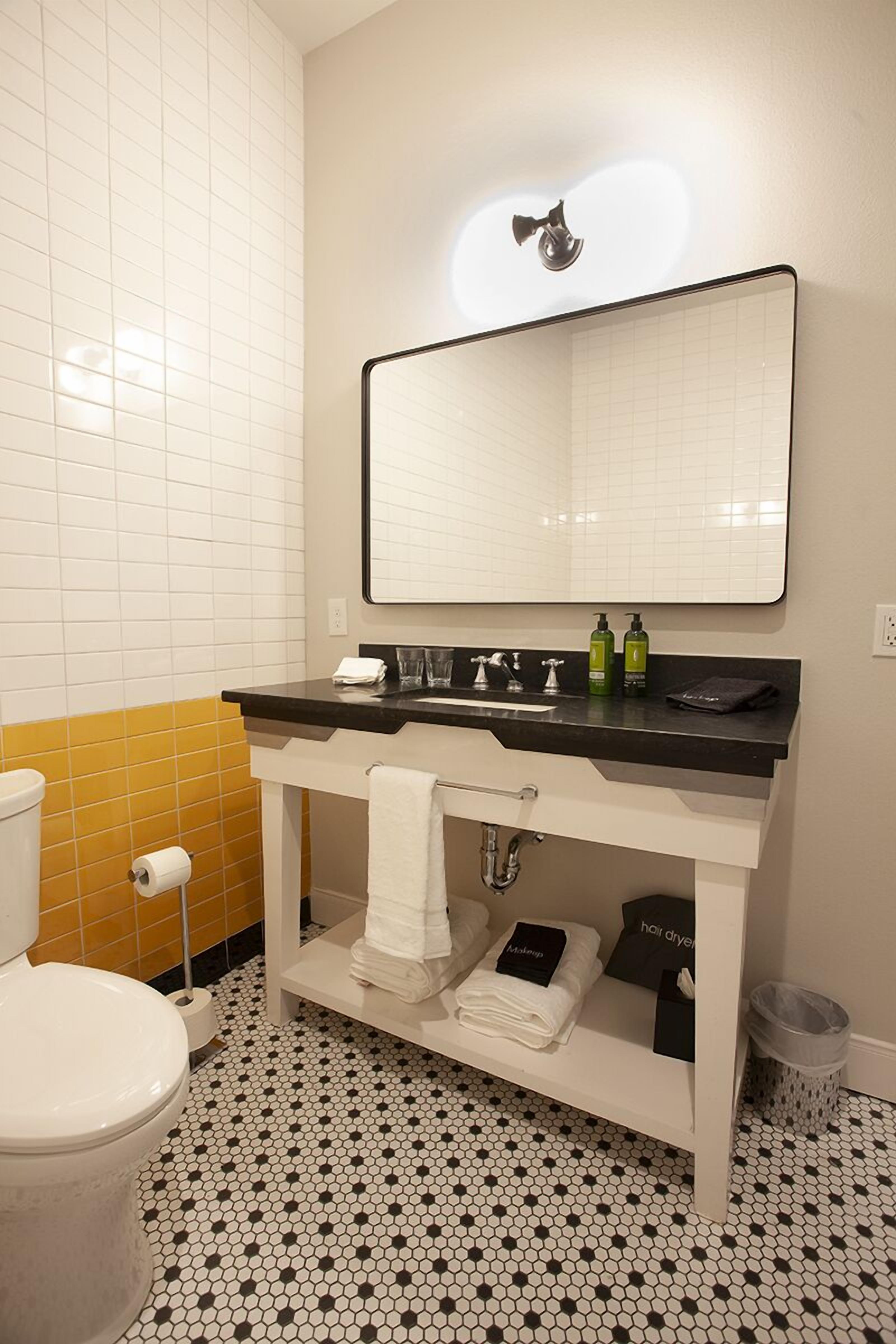 A bathroom with a white toilet, a black and white tiled floor, a black sink with a mirror above, and a yellow-tiled wall.