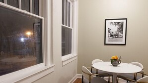 Un coin repas avec une table ronde et quatre chaises, un tableau encadré au mur et de grandes fenêtres avec des encadrements blancs.