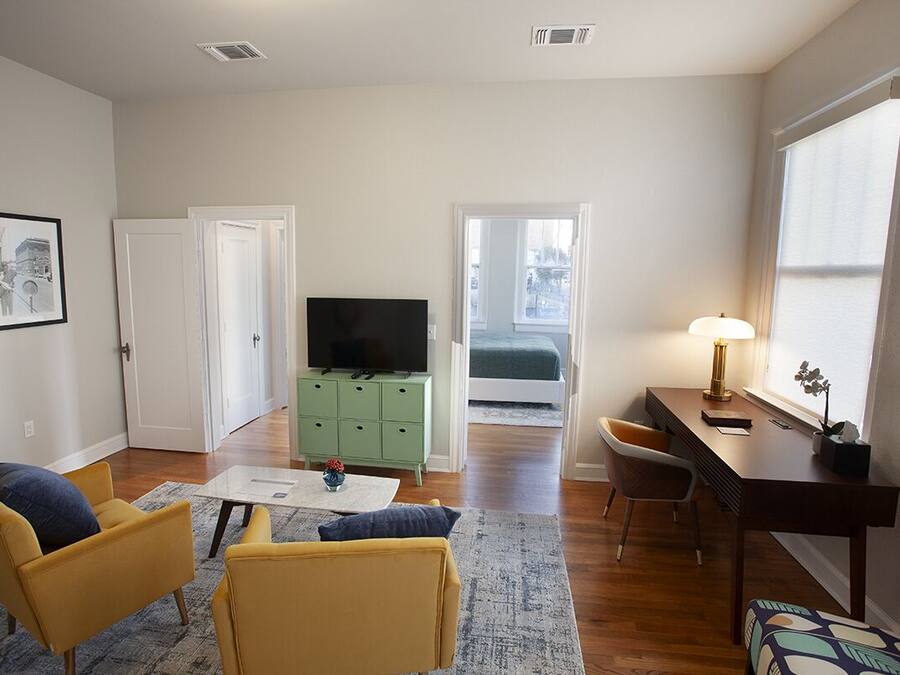 Una sala de estar con sofá, mesa de centro, televisor, escritorio y lámpara. Hay una puerta que da a un balcón.