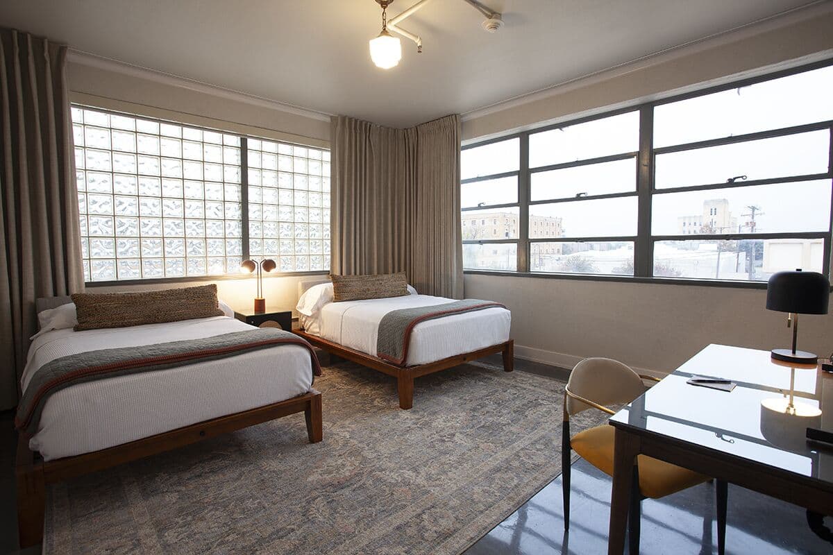 A hotel room with two beds, a desk, and large windows offering a city view.
