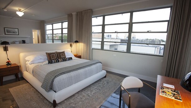 Une chambre d’hôtel moderne, dotée d’un grand lit, d’un bureau et offrant une vue sur la ville grâce à de grandes fenêtres.