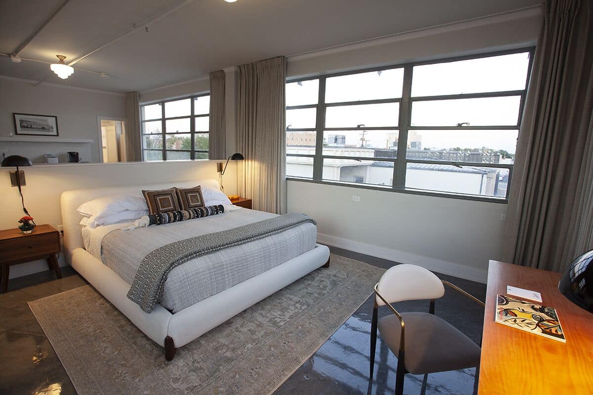 A modern hotel room with a large bed, a desk, and a view of the city through large windows.