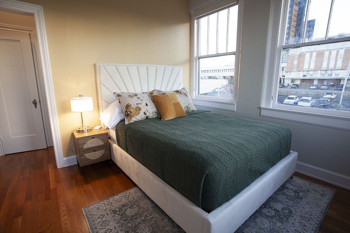 A bedroom with a bed, nightstand, lamp, and a view of the city through large windows.