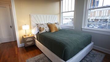 A bedroom with a bed, nightstand, lamp, and a view of the city through large windows.