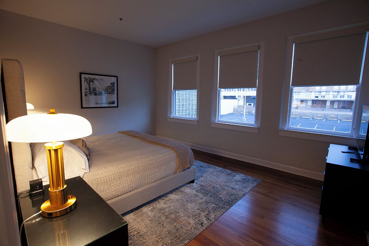 A bedroom with a bed, bedside lamp, windows, and a desk.