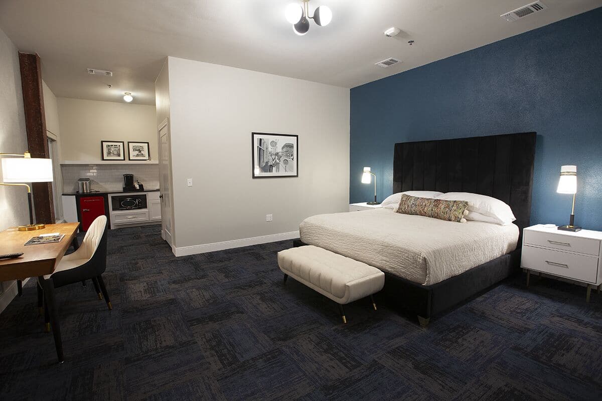 A hotel room with a bed, bedside tables, a desk with a chair, a kitchenette, and a bathroom.