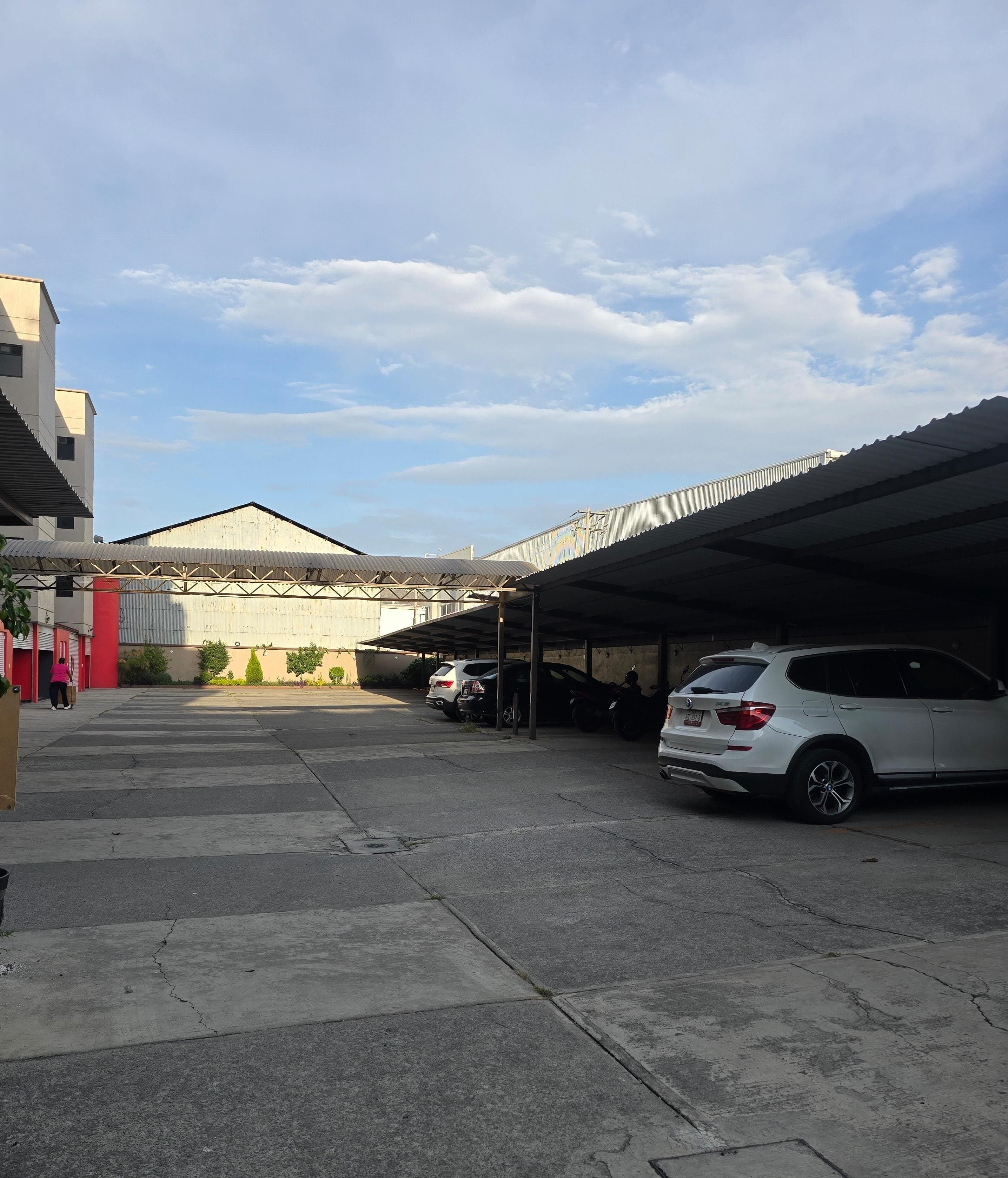 Un estacionamiento con estructuras cubiertas, una camioneta SUV blanca y un edificio con techo de metal corrugado.