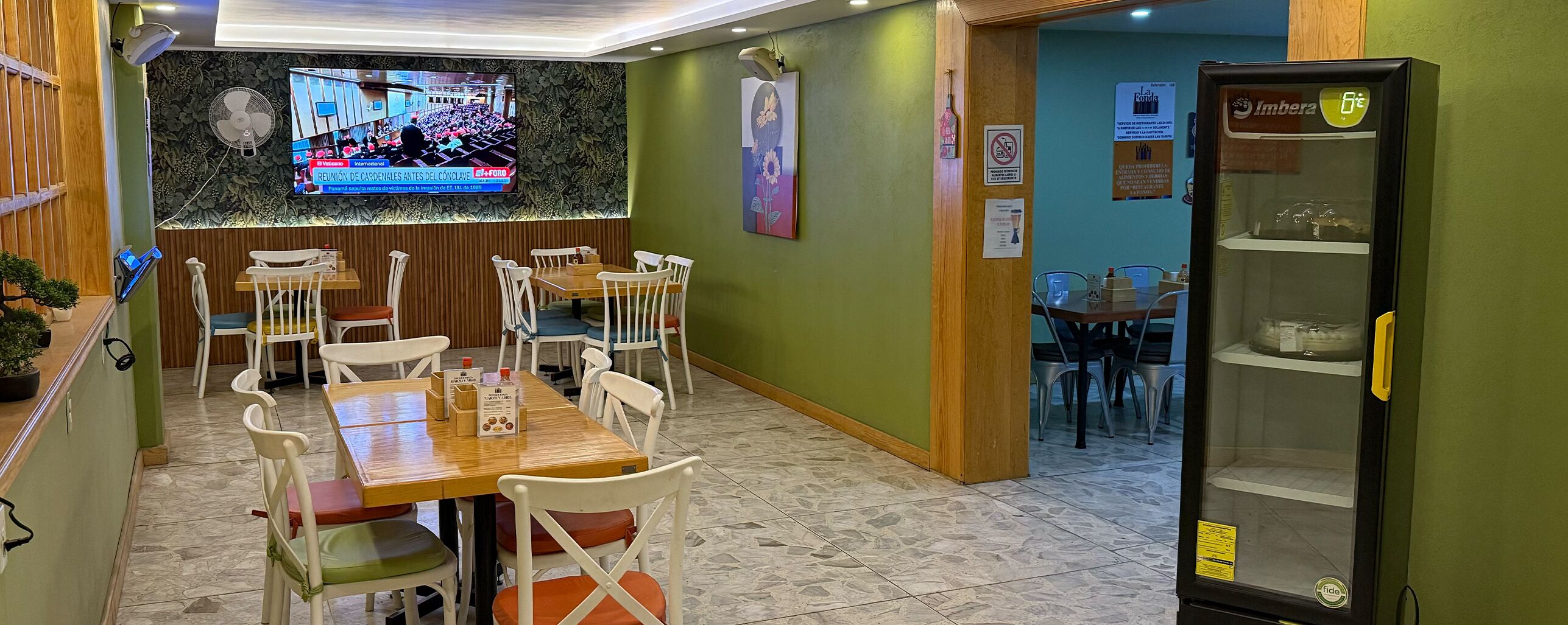 Ein Restaurant mit Fernseher, der eine Nachrichtensendung zeigt, Holztischen und -stühlen sowie einem Kühlschrank.