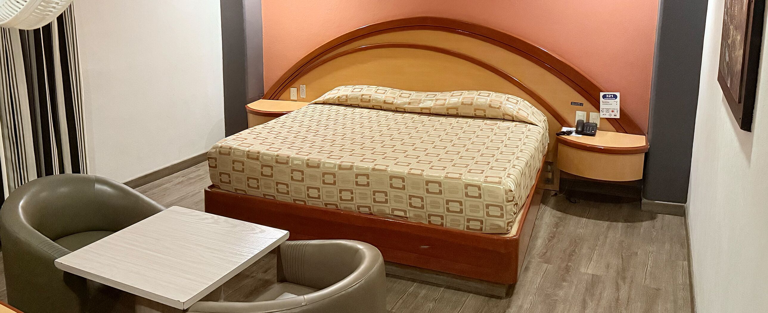 A hotel room with a bed, a chair, a small table, a nightstand, and a wall-mounted phone.