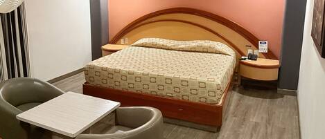 A hotel room with a bed, a chair, a small table, a nightstand, and a wall-mounted phone.