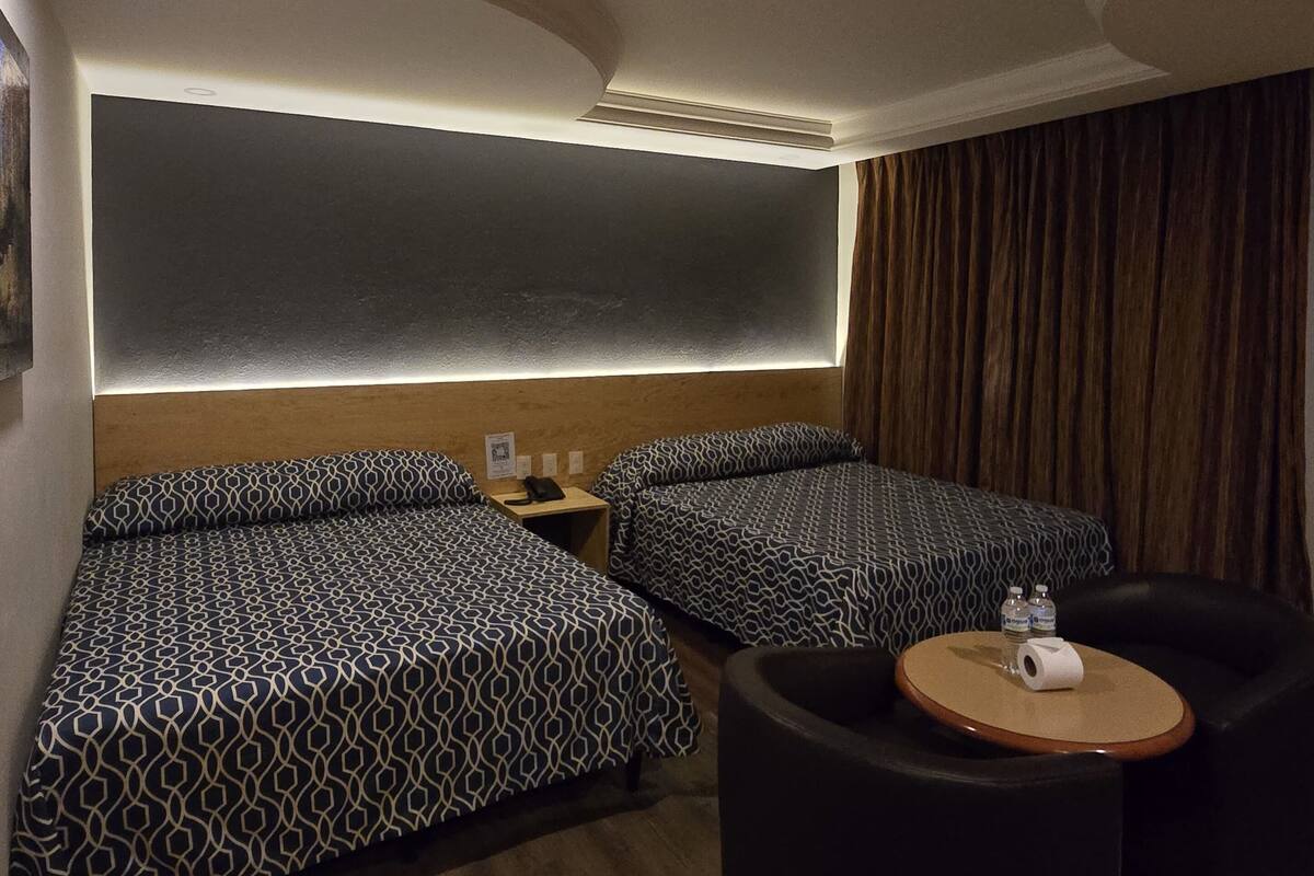 A hotel room with two beds, a small table with a chair, and a large window with curtains.