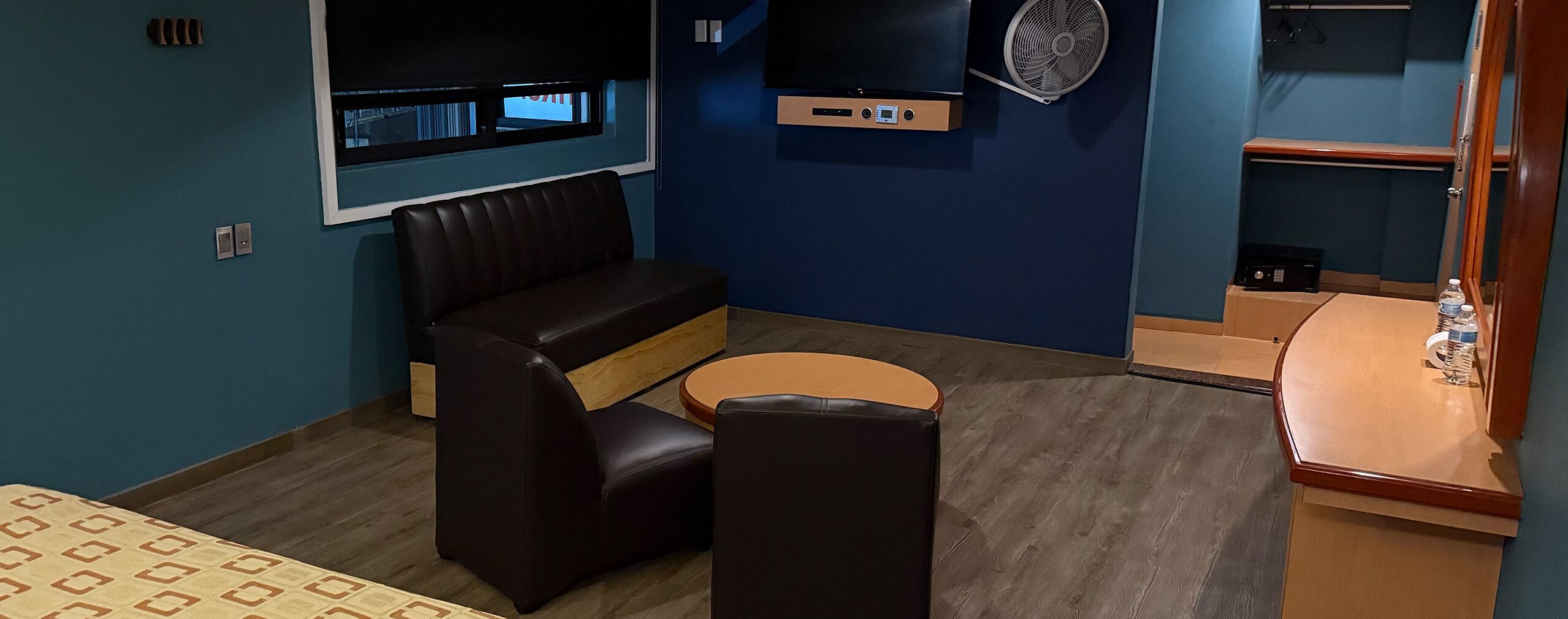 Una habitación con un sofá, una pequeña mesa redonda, un televisor montado en la pared y un ventilador.