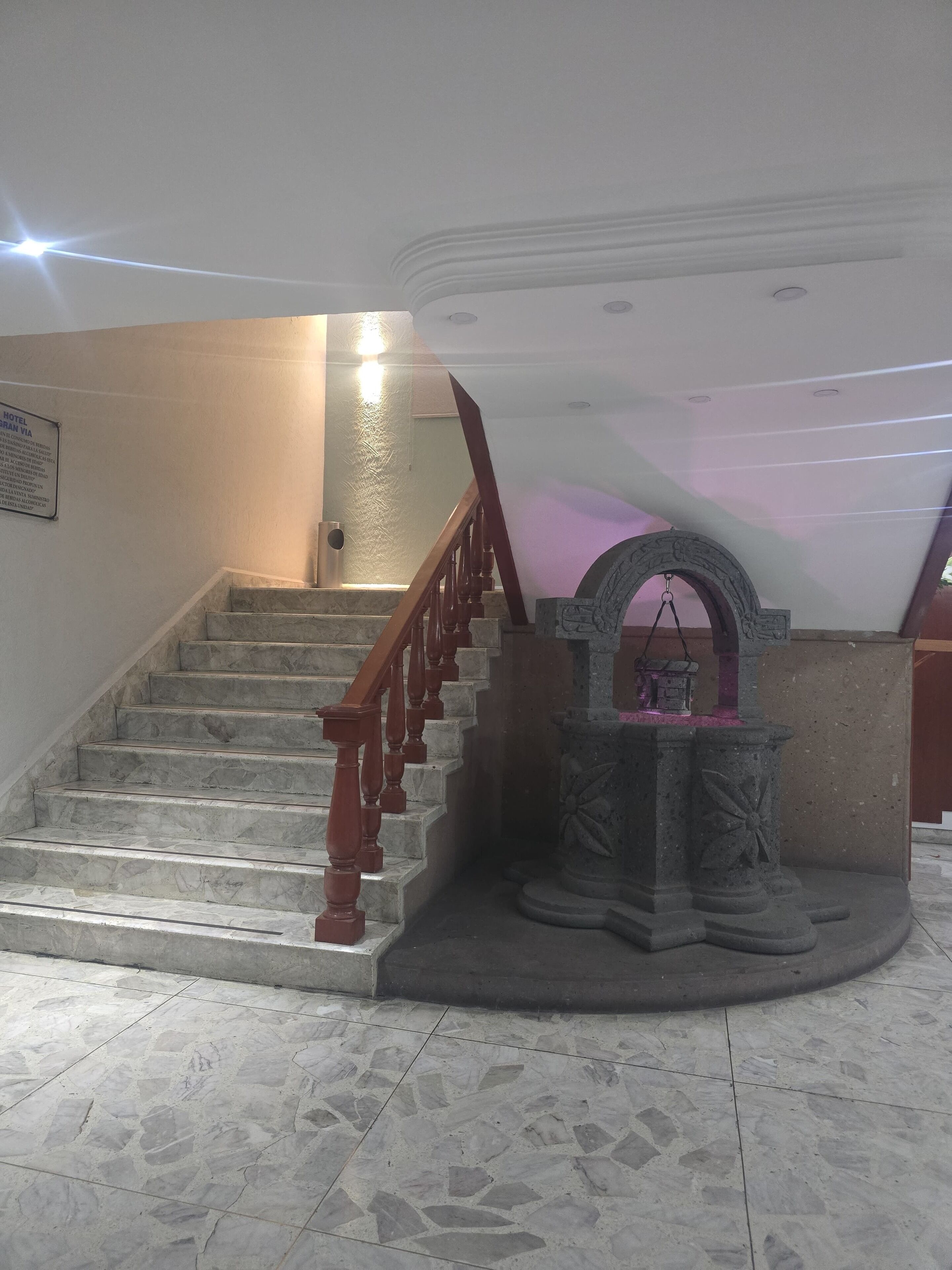 A staircase with marble steps and wooden railings, leading to a landing with a stone fountain and a wall-mounted sign.
