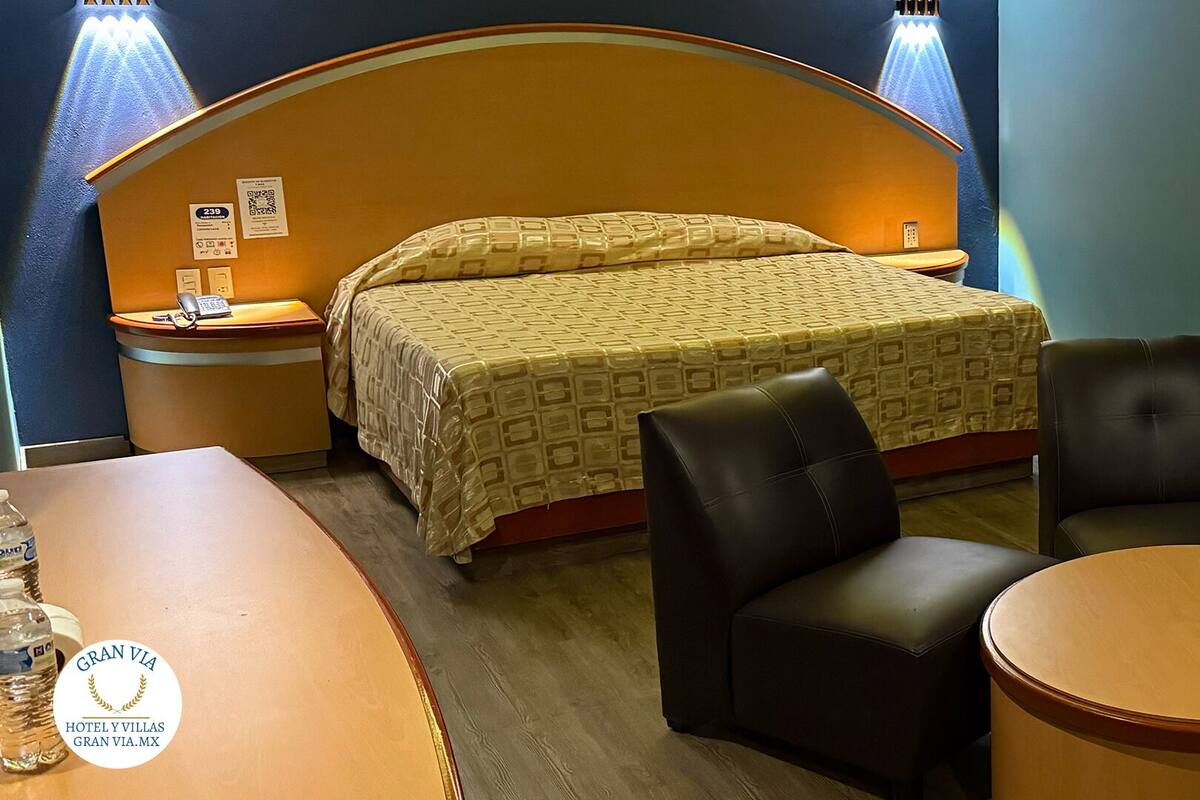 A hotel room with a bed, two chairs, a small table, a sofa, and a nightstand with a phone and a QR code.