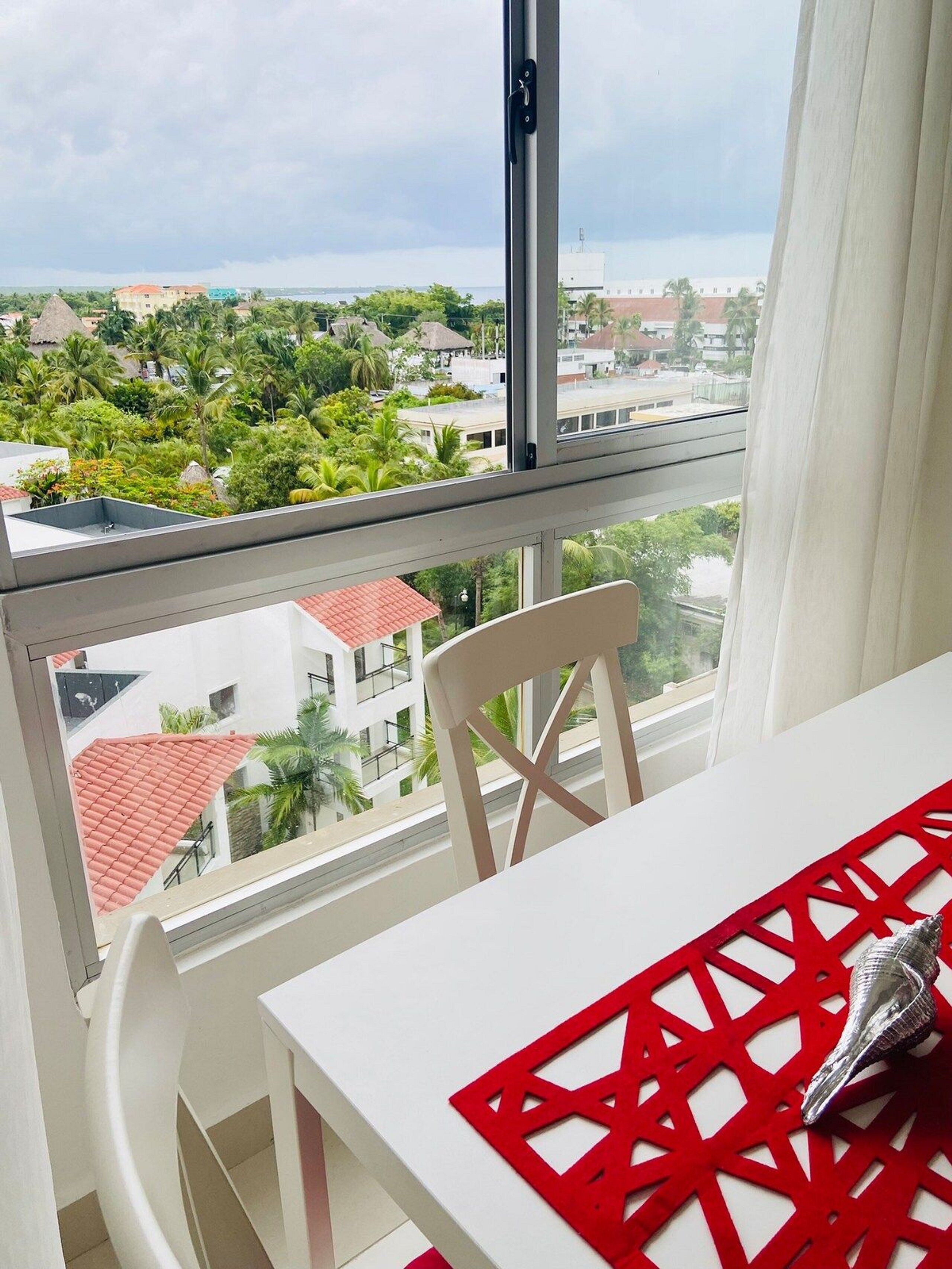 A balcony with a white table and chair, overlooking a residential area with red-roofed buildings and greenery.
