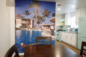 Un coin repas avec vue sur une piscine et des palmiers, une cuisine avec des meubles blancs et un réfrigérateur vert, ainsi qu’une table en bois avec des chaises.