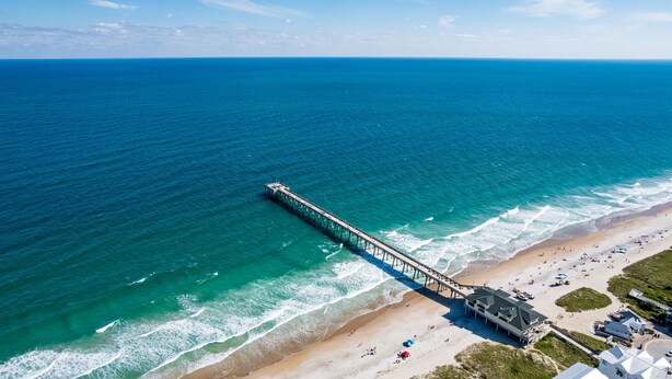 An aerial view of a beach with a pier extending into the ocean, a sandy beach, and a building with a sloped roof. - Coral House - Waterfront Beach Getaway, Stunning Sunsets, 9 min walk to beach! (Wrightsville Beach)
