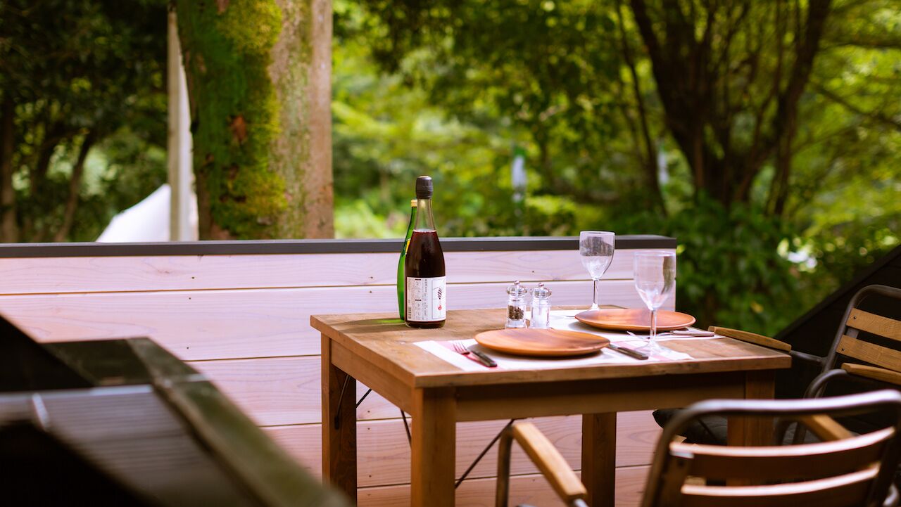 A wooden table set outdoors with a bottle of wine, glasses, and cutlery.