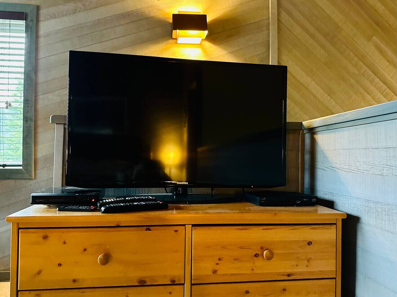 A television on a wooden dresser with a wall-mounted light above it.