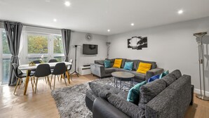 A modern living room with a grey sofa, a dining table with chairs, and a flat-screen TV mounted on the wall. - 3br-sleeps8-canarywharf-cinemabedroom (London)