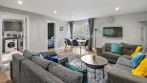 A modern living room with grey sofas, a coffee table, and a dining area with a table and chairs. There is a TV mounted on the wall and a washing machine in the corner. - 3br-sleeps8-canarywharf-cinemabedroom (London)