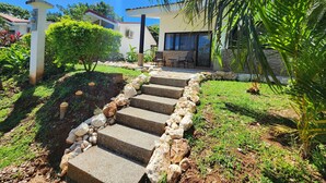 Eine Steintreppe führt zu einem Haus mit einer Terrasse, umgeben von Grünpflanzen und einem strahlend blauen Himmel.