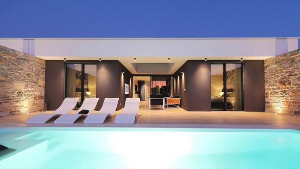 Un espace moderne aménagé au bord de la piscine, comprenant des chaises longues, un coin repas et une chambre visible à travers des portes vitrées.